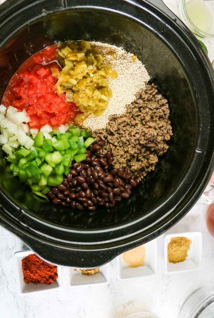 Slow cooker filled with raw quinoa, beans, ground beef, diced tomatoes, peppers, onions, and green chilies—everything you need for a hearty crockpot enchilada casserole.