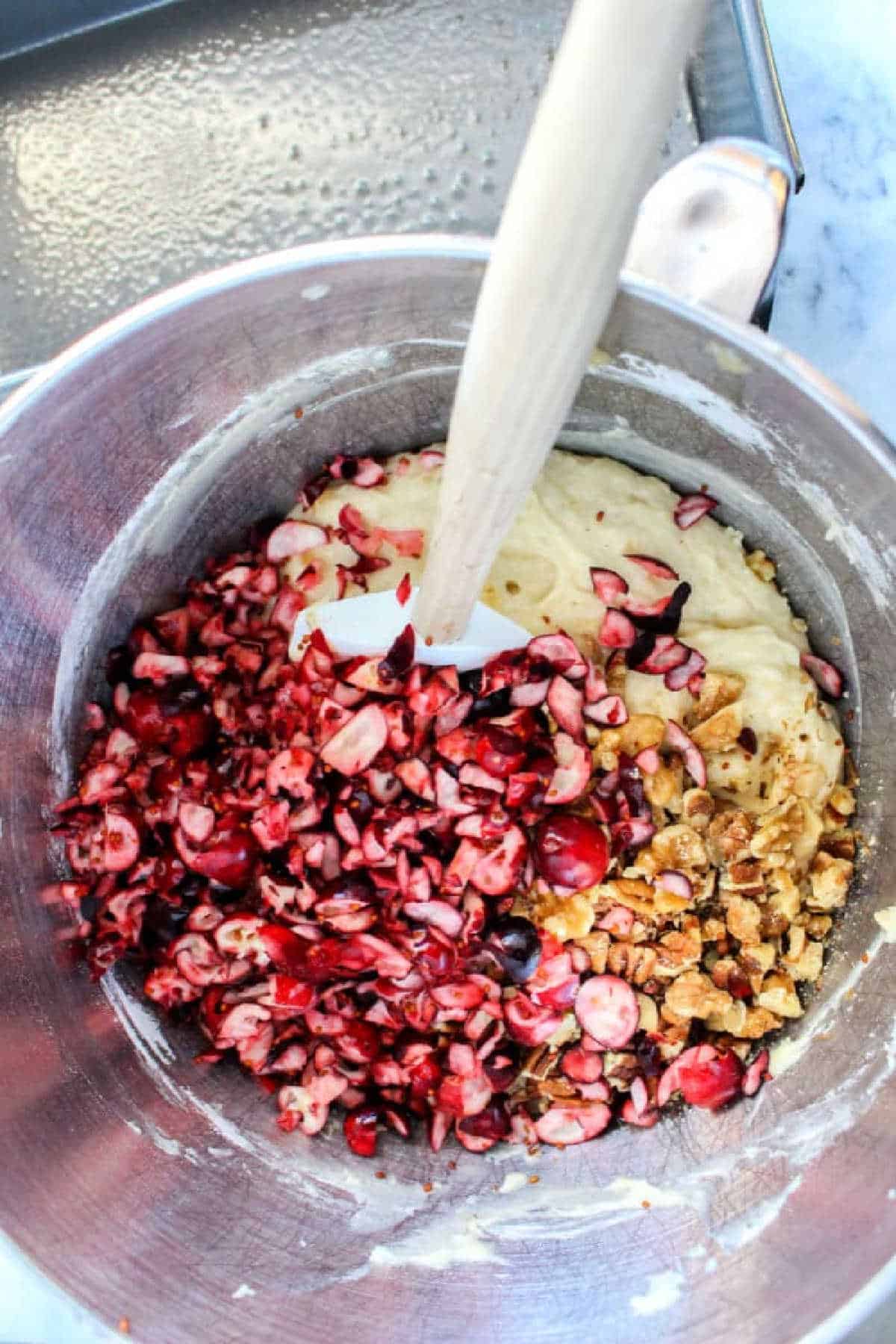 Chopped cranberries and walnuts are being mixed into batter for delicious banana cranberry bread in a metal bowl with a spatula.