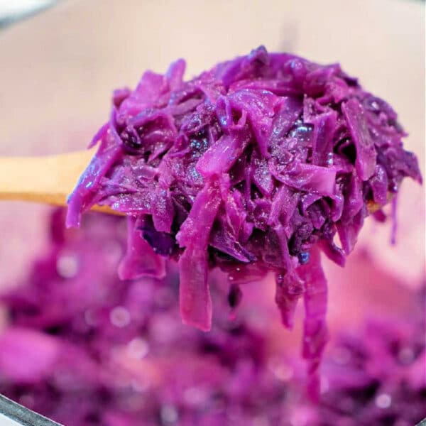 A wooden spoon holding sauteed red cabbage over a pot filled with more shredded cabbage.