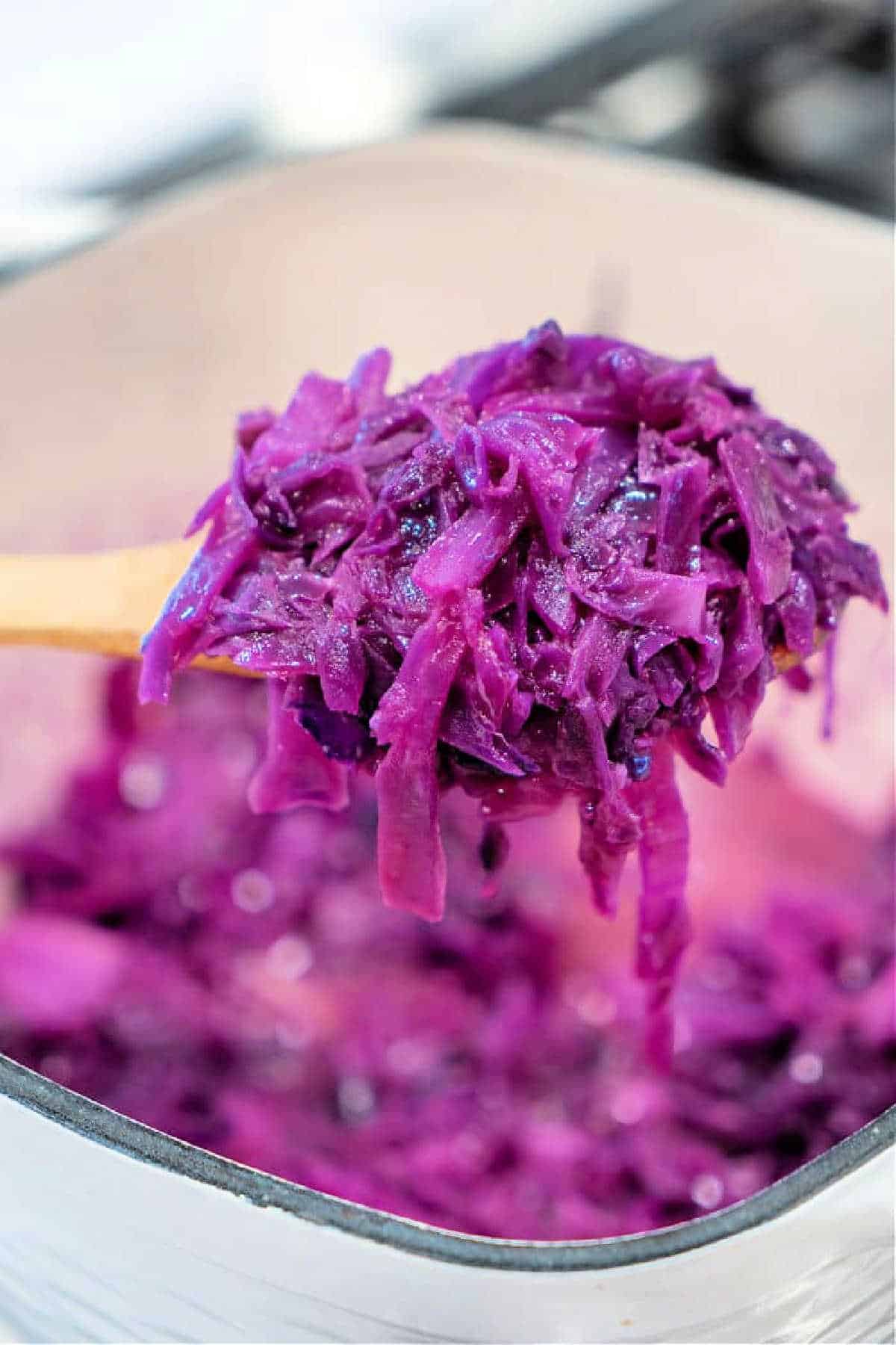 A wooden spoon holding sauteed red cabbage over a pot filled with more shredded cabbage.