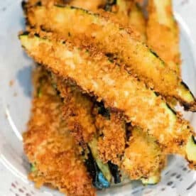 Golden, crispy air fryer zucchini fries stacked on a white plate, coated in breadcrumbs.