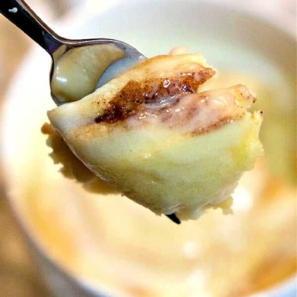 A spoonful of gooey cinnamon roll in a mug mug cake is held above a white mug.