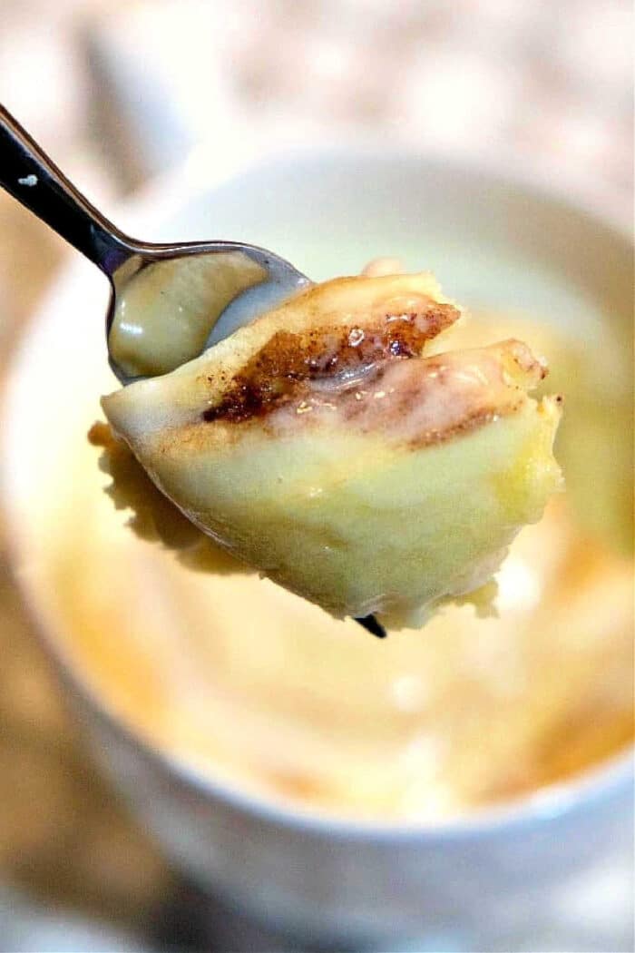 A spoonful of gooey cinnamon roll in a mug mug cake is held above a white mug.
