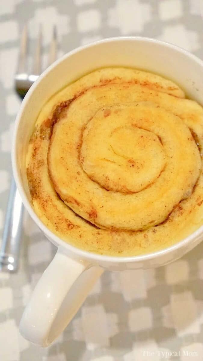 A cinnamon roll in a mug sits in a white cup with a fork beside it on a checkered surface.