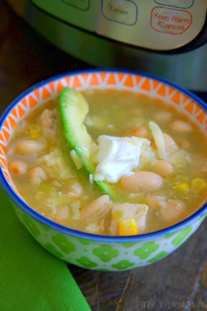 A bowl of instant pot white chicken chili topped with avocado and sour cream sits in front of an electric pressure cooker.
