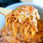 A wooden spoon lifting cheesy Ninja Foodi spaghetti from a pot, with melted cheese and herbs on top.