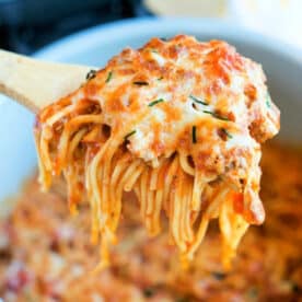 A wooden spoon lifting cheesy Ninja Foodi spaghetti from a pot, with melted cheese and herbs on top.