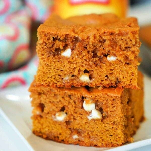 Two pumpkin blondies with white chocolate chips are stacked on a white plate, highlighting the delicious fusion of fall flavors and creamy sweetness.