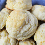 A close-up of freshly baked, golden-brown sour cream biscuits stacked on a blue cloth.