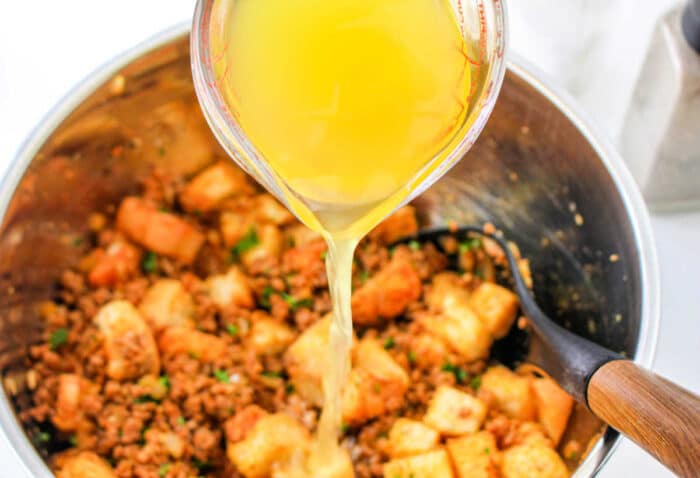 A measuring cup pours broth into a bowl of stuffing mix with bread cubes, sausage, and chorizo for a flavorful chorizo stuffing.