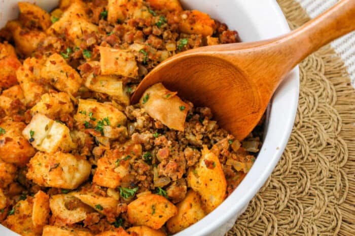 A wooden spoon scoops savory Chorizo Stuffing with bread cubes and herbs from a white dish.