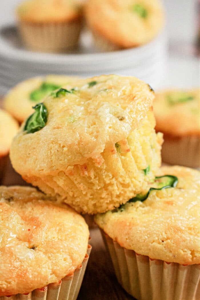 Close-up of Jiffy Corn Muffins topped with green herbs, stacked on a table with more corn muffins in the background—perfectly moist thanks to sour cream.