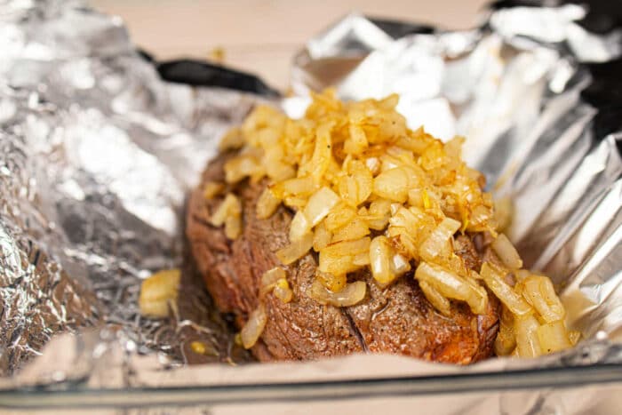 Juicy chuck roll steak cooked to perfection, topped with sautéed onions, and resting on foil in a glass baking dish.