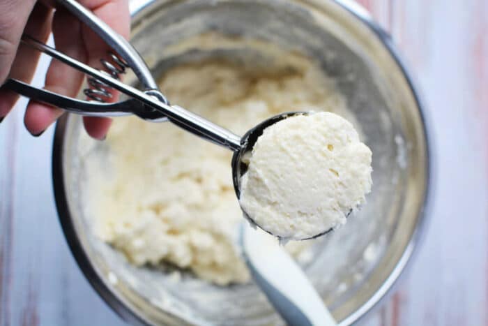 A hand holds a cookie scoop of dough over a mixing bowl with more dough and a spatula inside, perfect for preparing a classic Sour Cream Biscuits recipe.