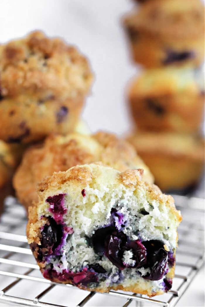 A zucchini blueberry muffin with a crumb topping sits on a cooling rack, with more muffins blurred in the background.