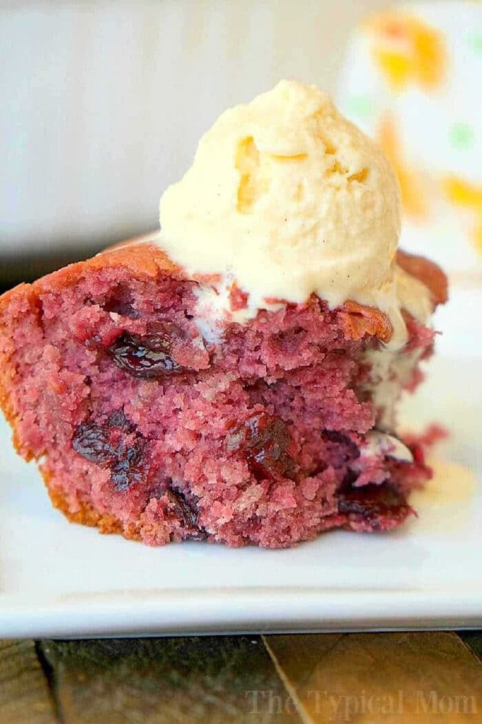 3 ingredient cherry pie filling dump cake for dessert! It's so simple to make even your kids can throw it together. It's really moist and packed with dark cherries or any other pie filling flavor you love. Cake mix baking has never been easier with this fun dump cake recipe. #cherry #pie #cake #dump #blueberry #easy #piefilling #cakemix #recipes A slice of berry cake made with cherry pie filling dump cake, topped with a scoop of vanilla ice cream on a white plate.