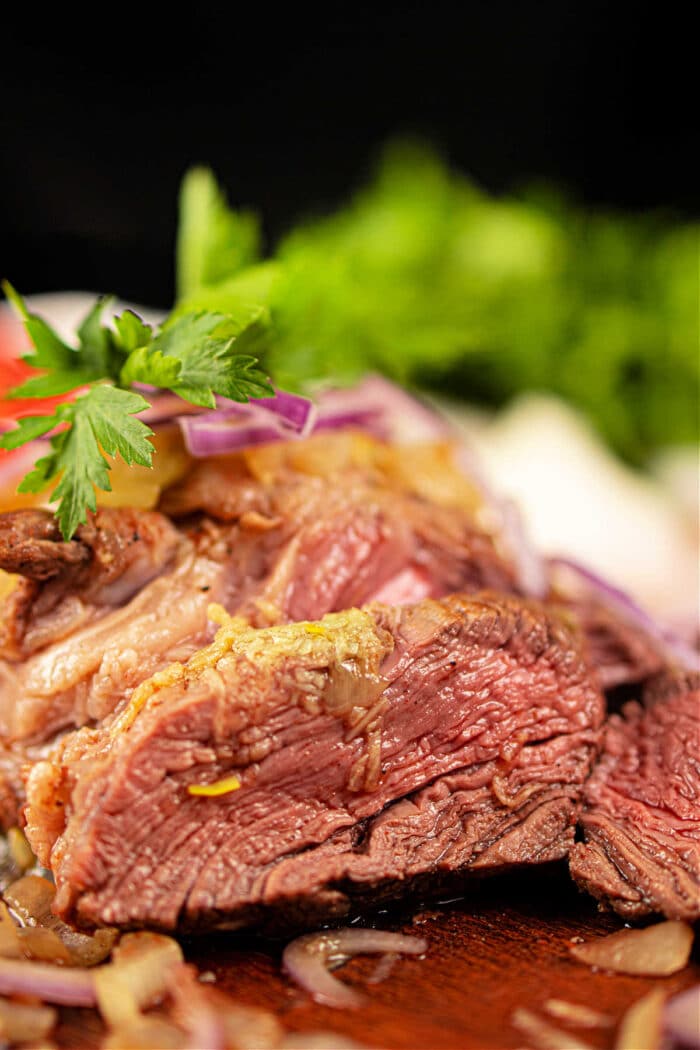 Close-up of juicy, medium-rare chuck roll steak garnished with parsley and sliced red onions, with fresh greens in the background.