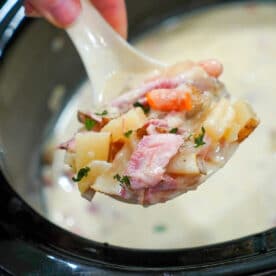 A hand holds a ladle of creamy soup with potatoes, carrots, and herbs over a slow cooker, serving up delicious crockpot ham and potato soup.