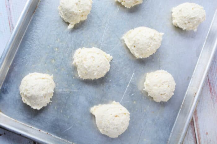 Scoops of raw Sour Cream Biscuits dough spaced on a metal baking sheet, ready to be baked.