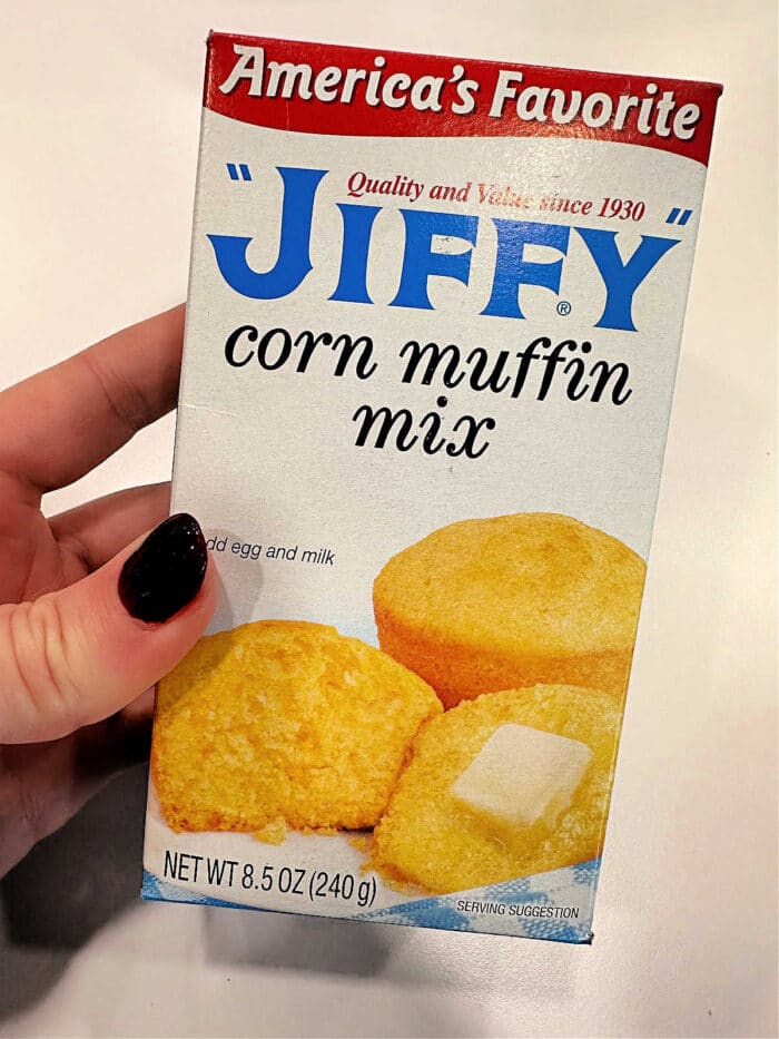 Hand holding a box of Jiffy Corn Muffins mix, featuring delicious muffins and a butter pat on the front—perfect for trying your favorite corn muffin recipe or adding sour cream for extra moisture.