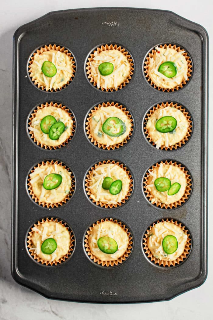 A muffin tin with 12 cups of unbaked Jiffy Corn Muffins, each topped with sliced jalapeños and loaded with cheesy goodness.