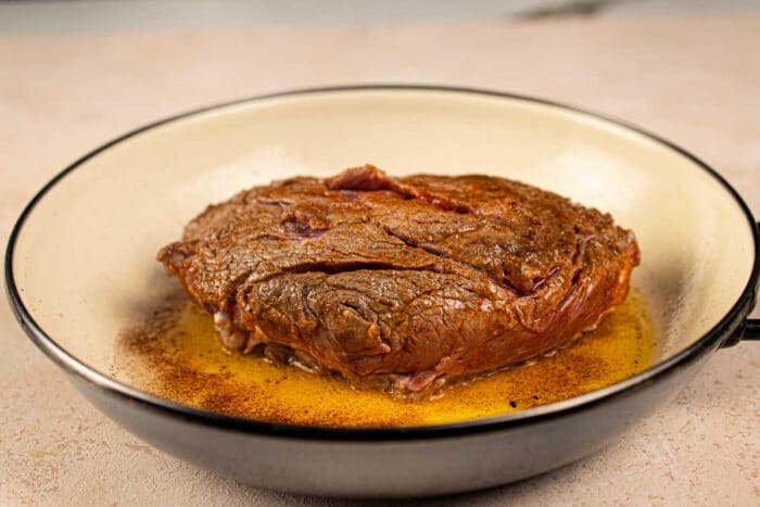 A seared chuck roll steak in a pan with sizzling juices and beautifully browned edges.