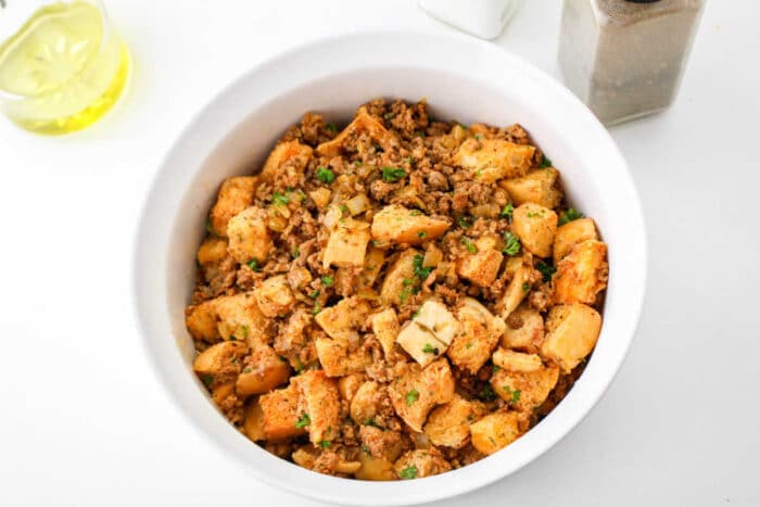 A bowl of homemade chorizo stuffing with bread cubes, herbs, and ground meat sits on a white surface.