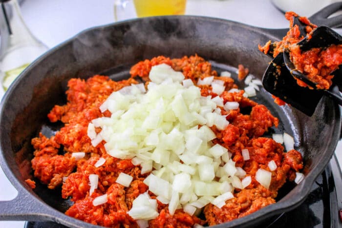 Chopped onions on top of raw ground meat in a black skillet, being stirred with a spatula—perfect as the base for a flavorful chorizo stuffing or your favorite stuffing recipe.