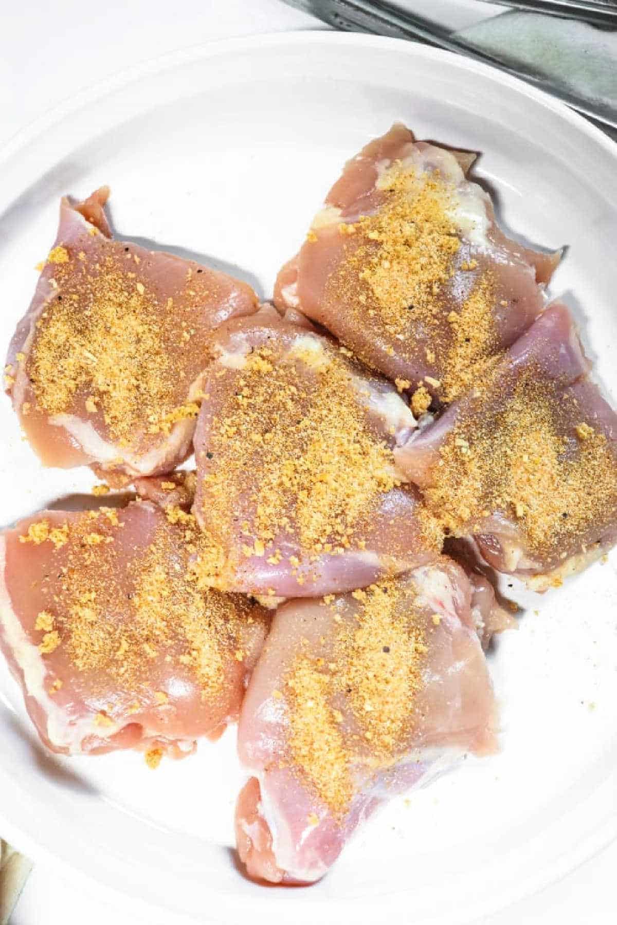 Raw chicken thighs on a white plate, sprinkled with a yellow seasoning powder—perfect for making tender Slow Cooker BBQ Chicken Thighs.