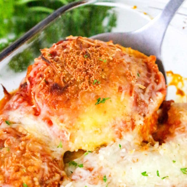 A serving of cheesy, baked eggplant parmesan is lifted from a glass dish, garnished with herbs—perfect alongside a comforting Ravioli Casserole.
