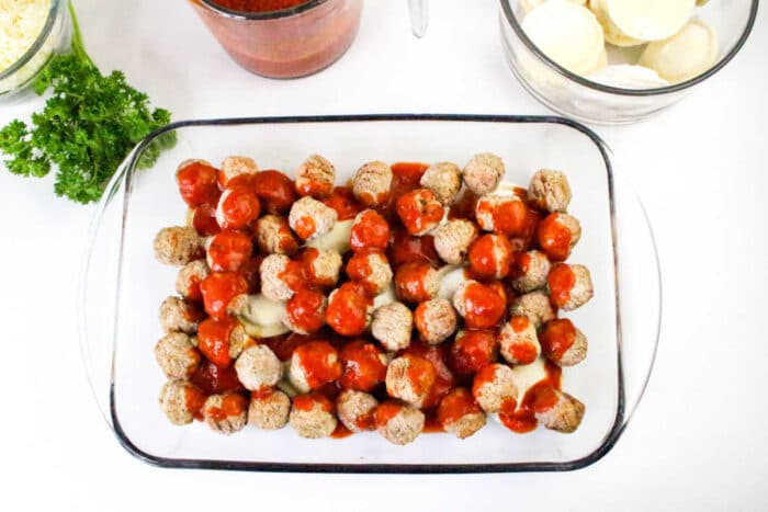 Glass baking dish with meatballs covered in tomato sauce, sliced mozzarella, and parsley on a white surface&mdash;perfect for serving alongside a homemade Ravioli Casserole.