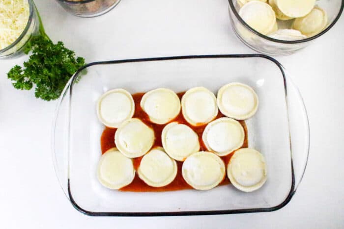 Glass baking dish filled with Ravioli Casserole on rich tomato sauce, surrounded by bowls and fresh parsley on a white surface.