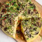 A sliced mushroom and herb frittata, inspired by an Instant Pot Quiche, sits on a wooden board with fresh chives sprinkled on top.