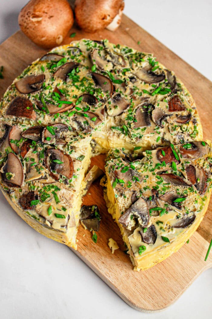 A sliced mushroom and herb frittata, inspired by an Instant Pot Quiche, sits on a wooden board with fresh chives sprinkled on top.