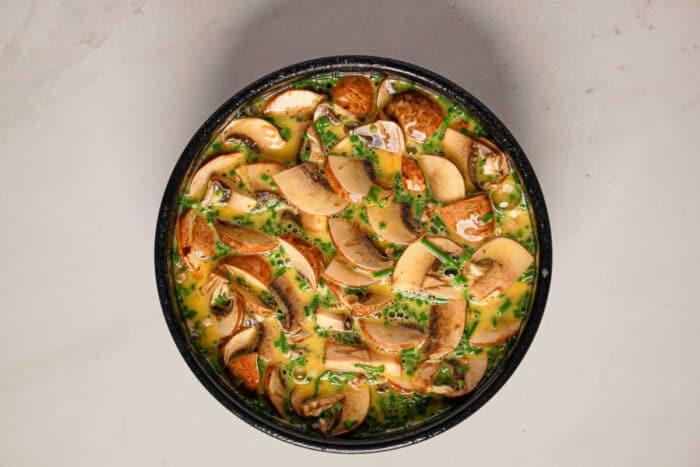A bowl filled with sliced mushrooms and chopped herbs in a beaten egg mixture, perfect for making a delicious Instant Pot Quiche, on a light surface.