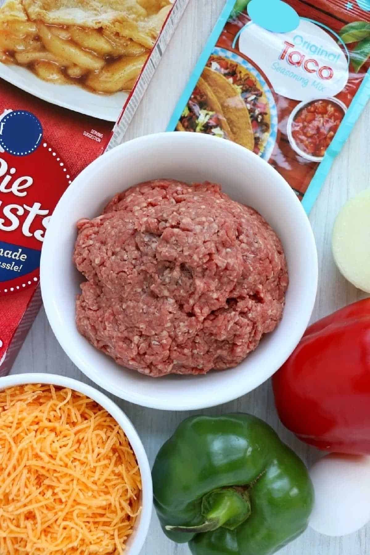 Ground beef in a white bowl with pie crusts, taco seasoning, cheese, onion, and peppers nearby—perfect for making air fryer empanadas at home.