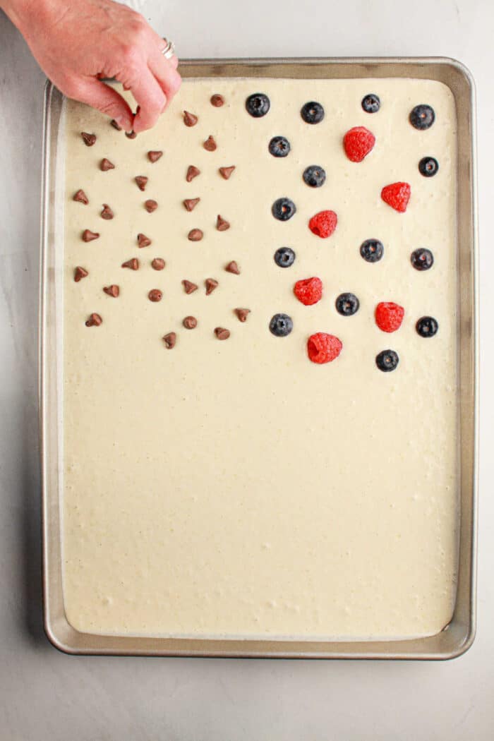 A hand decorates oven baked pancakes with pancake mix in a tray, adding chocolate chips, blueberries, and strawberries in a flag pattern.