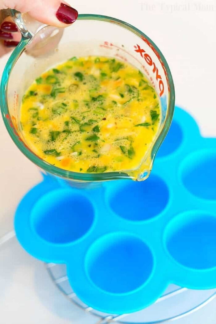 Pressure cooker egg bites are the perfect protein packed breakfast for busy mornings. A great Keto Instant Pot recipe with just a few ingredients. #instantpot #eggbites #starbucks #keto #instantpotrecipes #pressurecooker #crockpotexpress #thetypicalmom A hand pours egg mixture with vegetables from a measuring cup into a blue silicone mold to make pressure cooker egg bites.