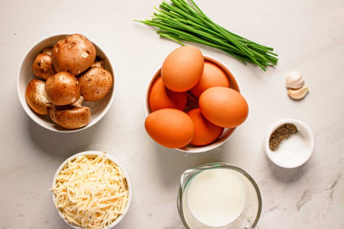 Brown eggs, mushrooms, shredded cheese, milk, chives, garlic, salt, and pepper arranged on a white surface—perfect ingredients for a homemade quiche or an easy Instant Pot breakfast.