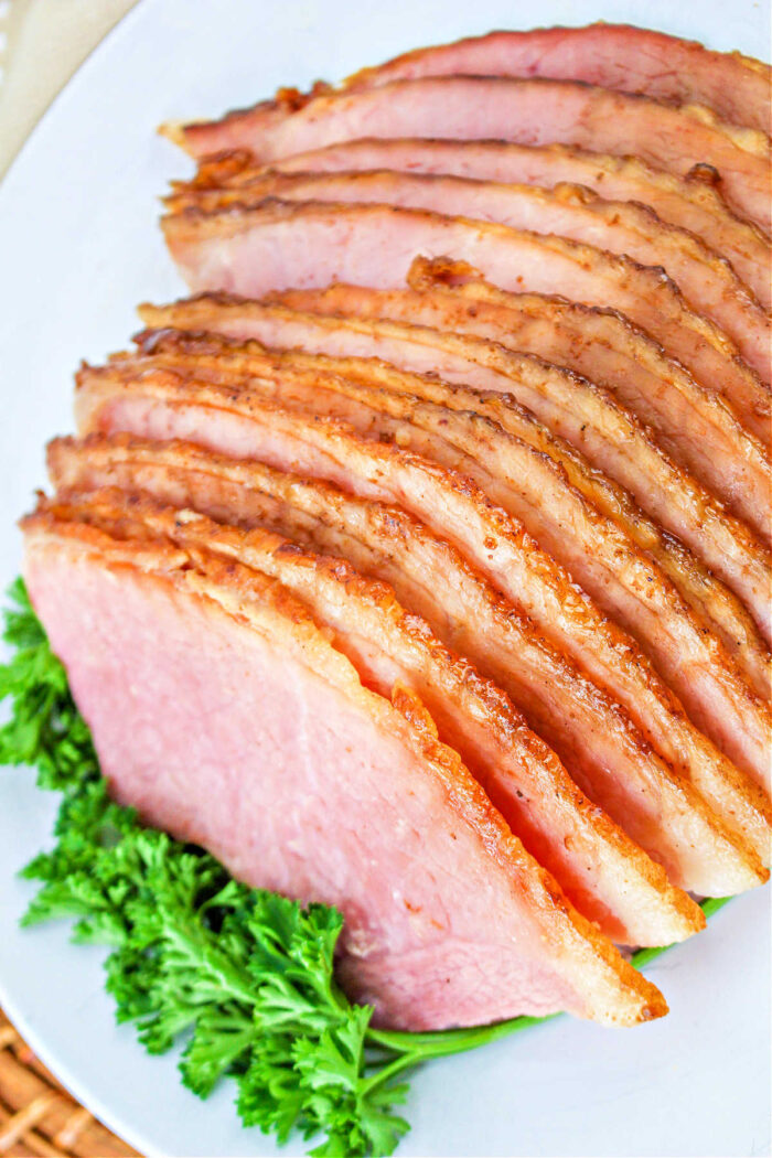 Sliced baked ham, prepared using the How to Cook a Spiral Ham in the Oven method, is elegantly arranged on a white plate with fresh parsley garnish.