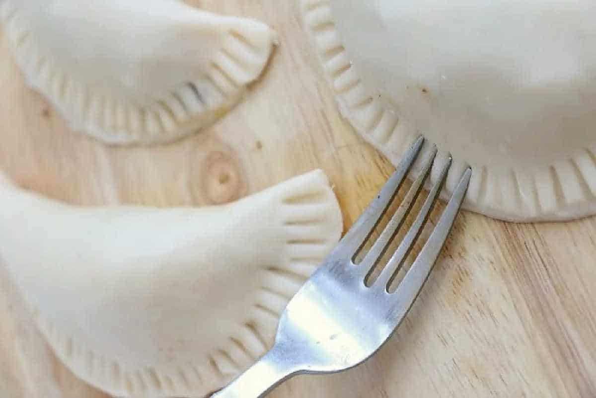 A fork pressing edges of uncooked air fryer empanadas on a wooden surface.