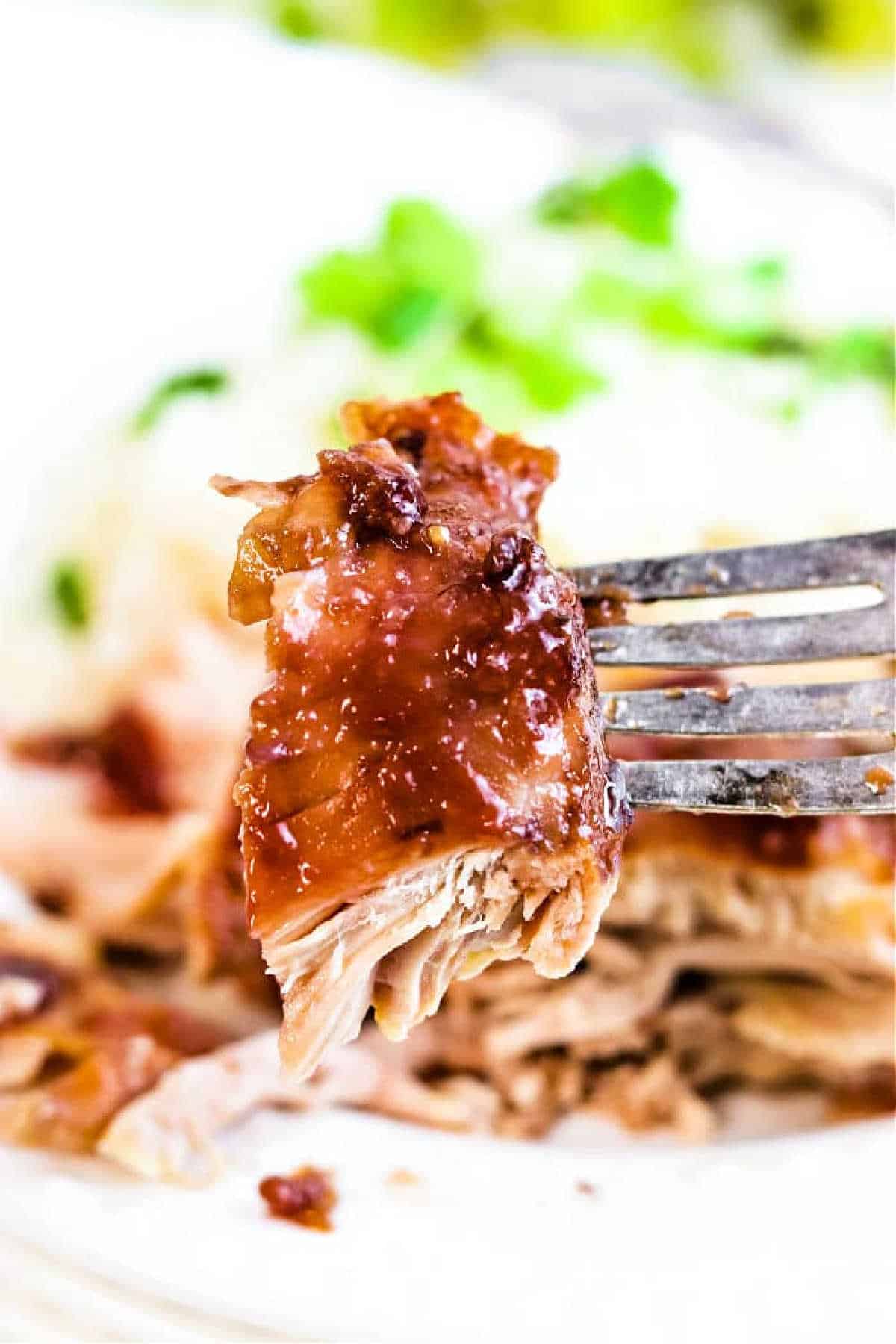 A close-up of a fork holding a juicy piece of glazed, shredded Slow Cooker BBQ Chicken Thighs with blurred greens in the background.