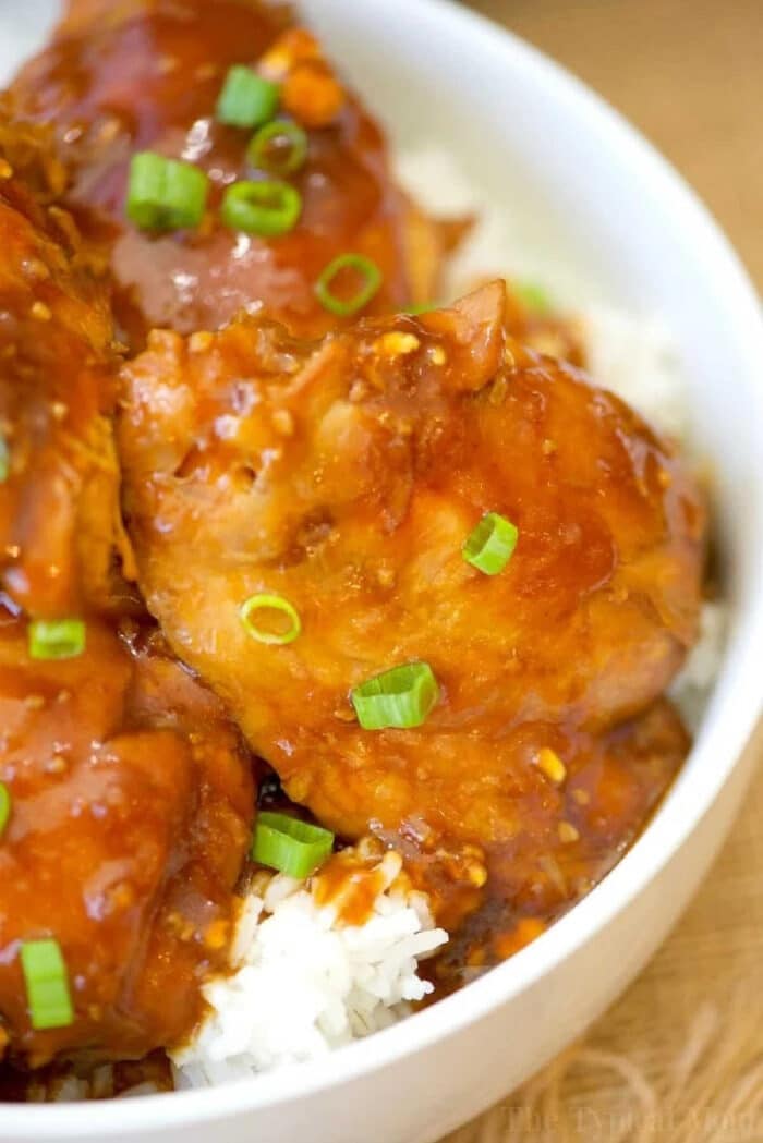 Instant Pot spicy teriyaki chicken thighs in a sticky orange glaze are served over white rice and garnished with chopped green onions in a bowl.