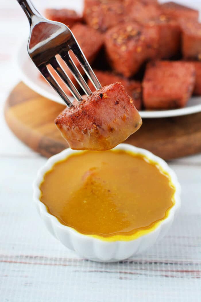 A fork holds a cube of Bologna Burnt Ends over a small bowl of mustard sauce, with more sausage pieces in the background.