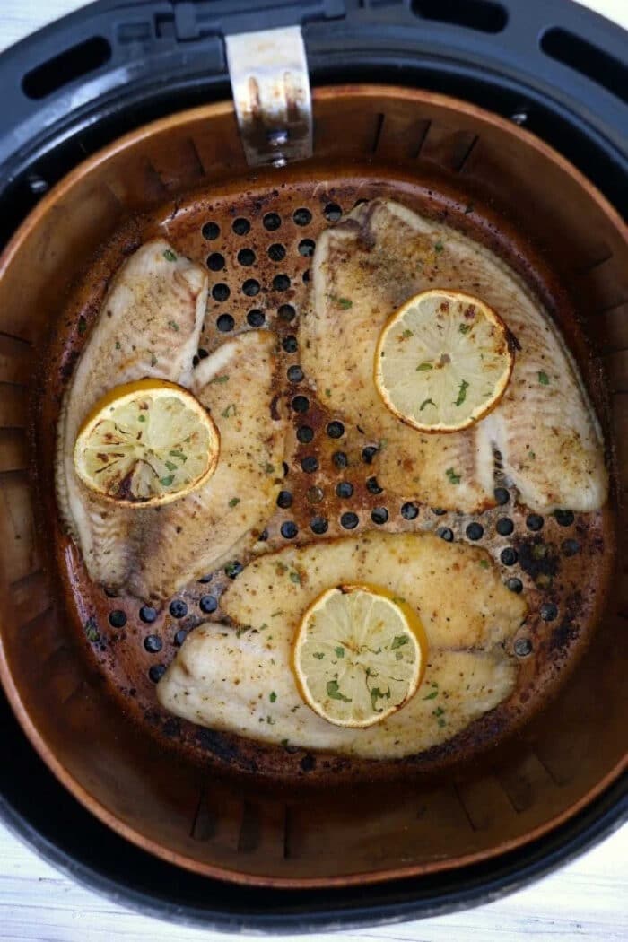 Four seasoned frozen fish fillets with lemon slices on top cooking in an air fryer basket.