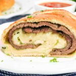 A sliced Philly Cheesesteak Stromboli with layers of beef and cheese on a white plate, sauce in the background.