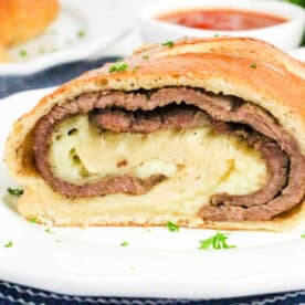 A sliced Philly Cheesesteak Stromboli with layers of beef and cheese on a white plate, sauce in the background.