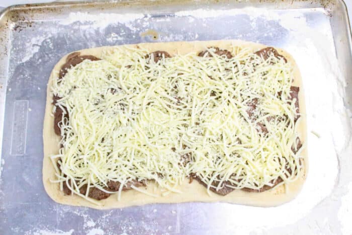 Rectangular pizza dough topped with meat slices and shredded cheese on a floured baking sheet, perfect for making Philly Cheesesteak Stromboli.