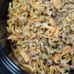 Close-up of creamy Ground Beef Stroganoff noodle casserole with herbs and crispy onions in a black slow cooker.