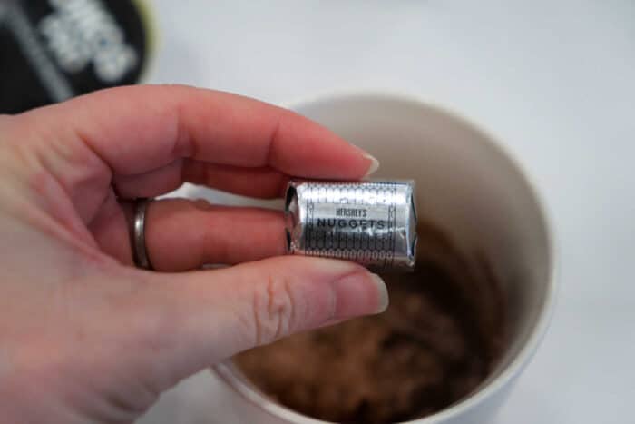 A hand holding a foil-wrapped Hershey’s Nuggets chocolate above a cup of hot cocoa, perfect for pairing with a slice of homemade chocolate banana bread.